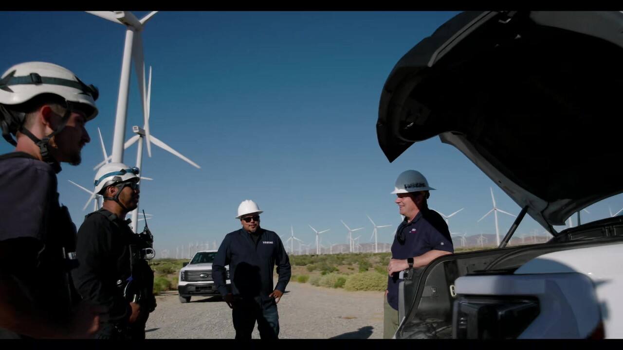 Four men wearing white construction hats stand in a circle outdoors, talking. Windmills can be seen spanning the grassy landscape in the background.